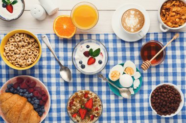Farklı kaseler ve bardaklar kareli bir kumaşa dizilmiştir. Masada mısır gevreği, yoğurt, meyve ve içecek var. Çeşitli yiyecek ve lezzetlerle hazırlanmış sıradan bir kahvaltı ortamı..