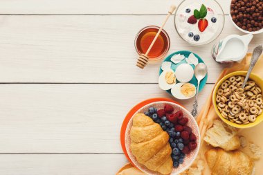 Bir sabah ziyafetinde kruvasan, yumurta, yoğurt, böğürtlen ve tahıl hafif ahşap bir masa üzerinde sıralanmış. Bal ve süt kahvaltı sahnesine çeşitli tabak ve kâselerle eklenir..