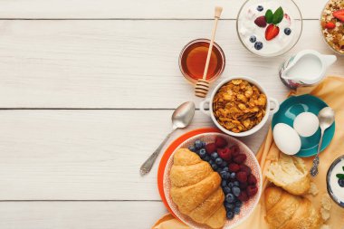 Kahvaltı yiyeceklerinin bir koleksiyonu ahşap bir masada düzenlenir. Kruvasan, meyve, yoğurt, bal ve daha fazlası var. Farklı dokular ve renkler, sabah yemeği için davetkar bir sahne yaratır..