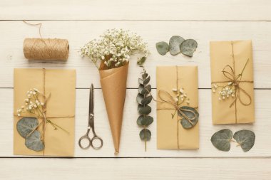 Kahverengi kağıtla paketlenmiş hediyeler ahşap bir masanın üzerine konur. Makas, sicim ve bir buket çiçek var. Okaliptüs yaprakları ve bebek nefesi çiçekleri dekorasyon için kullanılır..