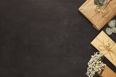 Hediye eşyalar, paketlerin ve zarfların gösterildiği karanlık bir yüzeye yerleştirilir. Kurumuş çiçekler ve okaliptüs gibi doğal elementler sahneyi tamamlayarak hediyeler için dekoratif bir ortam oluşturuyor..