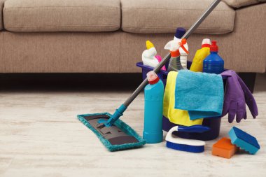 Bir kanepenin yanında bir temizlik malzemesi seti düzenlenir. Şişeler, süngerler ve bir paspas. Sahne temizlik için hazırlık gösteriyor..