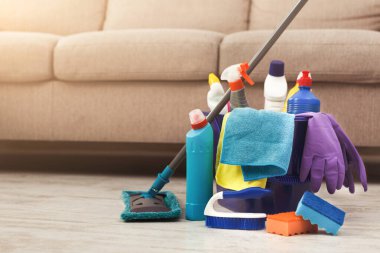 Temizlik malzemeleri oturma odasının zeminine bir kanepenin yanına konur. Farklı şişeler, elbiseler ve aletler gösteriliyor, temizlik işleri için hazır. Olay yeri parlak ve düzenli..