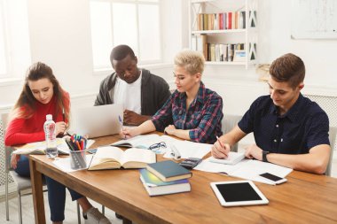 Dört öğrenci kitap ve malzemelerle dolu bir masanın etrafında toplandı. İşlerine odaklanmış, tartışıyor ve not alıyorlar. Alanın kitaplarla dolu geniş pencereleri ve rafları var..