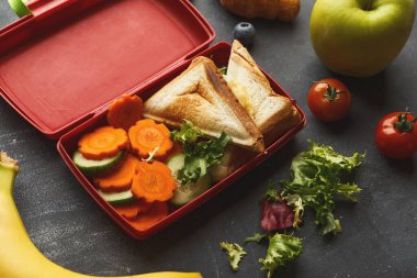 Kırmızı bir beslenme çantasında sandviçler, havuç dilimleri, salatalık ve marul var. Elma, muz ve domates gibi meyveler etrafa saçılmış. Öğlen yemeği vakti ve yemek hazır..
