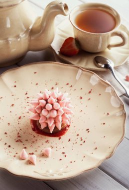 Sıcak bir fincan çay tabakta bir tatlının yanında oturur. Tatlı, çiçekleri andıran pembe dekoratif parçalardan oluşuyor. Ayar, sıcak bir atmosfer oluşturan basit sofra takımı ve elementlere sahip..