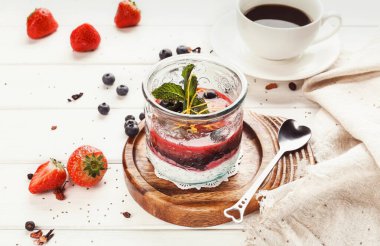 Bir tatlı ahşap bir tahtaya konur. Tatlı, meyve, krema ve nane katmanlarını içeriyor. Yakınlarda taze çilek ve yabanmersini var. Masanın üzerinde bir fincan kahve duruyor..