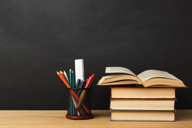 Bir dizi kitap ahşap bir masanın üzerinde duruyor. En üstte açık bir kitap var. Siyah bir arkaplan karşısında renkli kalemler, kalemler ve bir cetvel bulunan bir kap var..