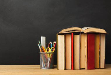 Kalem, kalem ve makasla dolu bir konteynırın yanındaki ahşap masada bir kitap seti duruyor. Arka plan karanlık bir yüzey. Bu sahne çalışma ya da çalışma alanı kurulumunu öneriyor.