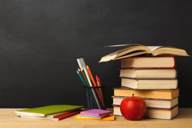 Bir kitap koleksiyonu parlak kırmızı bir elmanın yanında bir masanın üzerine istiflenmiştir. Kalemler ve kalemler bir kutuya konur. Not defterleri ve renkli kağıtlar var. Okul ortamını gösteriyor..