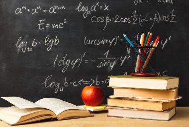 Bir masa istiflenmiş kitaplar, bir elma ve bir fincan renkli kalemlerle dolu. Arkalarında, bir karatahta çeşitli matematik denklemlerini gösteriyor. Okul ortamı..