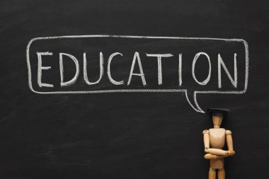 Tahta bir figür, eğitim sözcüğünün büyük harflerle yazıldığı bir tahtaya karşı duruyor. Sahne, öğrenme ve öğretme kavramlarına odaklanma anını yakalıyor..