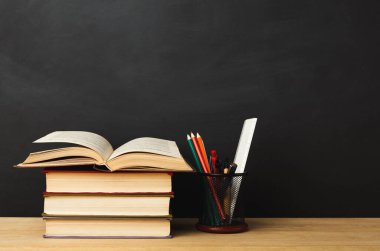 Masaya bir yığın kitap konur. Bir kitap açık, sayfalarını gösteriyor. Yakınlarda, bir kupada renkli kalemler ve kalemler var. Arka plan karanlık ve basit..