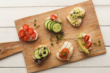 Farklı küçük sandviçler tahta kesme tahtasının üzerinde oturur. Her sandviçte domates, salatalık ve yeşillik gibi çeşitli malzemeler var. Bu sahne paylaşıma hazır rengarenk abur cuburları gösteriyor..