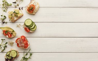 Açık yüzlü sandviçler ahşap bir masada düzgünce düzenlenir. Her sandviçin farklı sosları var. Dilimlenmiş salatalık, domates ve otlar da dahil. Taze malzemeler görünür..
