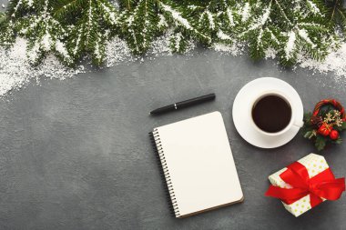 A collection of winter items lays on a gray surface. There is a cup of coffee, a blank notebook, a gift wrapped in colorful paper, and green decorations. Snow-like items are scattered around.