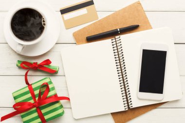 A collection of items on a table shows a cup of coffee, a blank notebook, a smartphone, and a gift box. These items suggest preparing for events or activities. A credit card lies nearby.