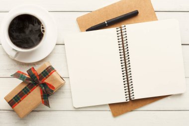 A collection of items including a cup of coffee, a wrapped gift, and an open notebook with a pen sits on a wooden table. This scene captures a moment of preparation for writing or planning.