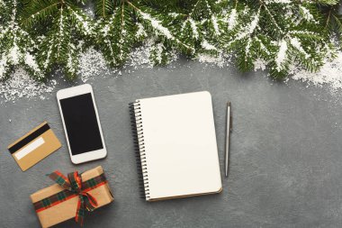 Bir defter, akıllı telefon, hediye kutusu ve hediye kartı da dahil olmak üzere çeşitli eşyalar gri bir yüzeyde sıralanır. Çam dalları ve kar kış temasına ilave ederek, tatillere hazırlanıyor..