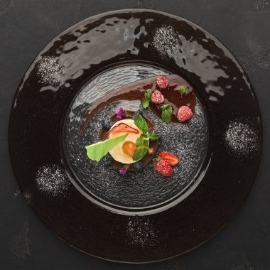 Bir tatlı koleksiyonu siyah bir tabakta düzenlenir. Tabakta krema, meyve ve yeşil otlar yer alıyor. Çekici bir görünüm yaratan farklı dokular ve renkler sergiliyor.