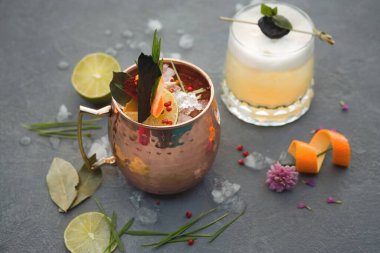 Gri yüzeyde iki farklı kokteyl sunulur. Bir kokteyl bakır bardakta, buzla dolu renkli otlar ve meyvelerle süslenmiş..