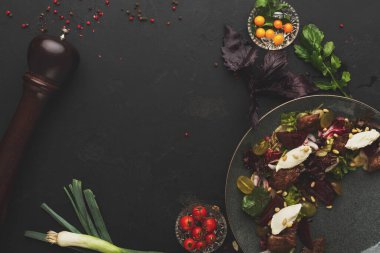 Taze salata malzemelerinden oluşan bir koleksiyon karanlık bir yüzeye yerleştirilir. Tabakta sebzeler, domatesler ve baharatlar yer alıyor. Çeşitli kaselerde otlar gibi ilave malzemeler bulunur..