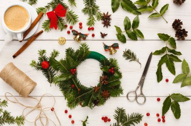 Tatil çelengi yapmak için bir malzeme koleksiyonu masaya yayılır. Yeşil yapraklar, kırmızı böğürtlen, kozalak, makas, ip ve yakınlarda bir fincan kahve var..