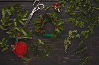 Tahta bir yüzeyde çelenk yapmak için bir malzeme koleksiyonu sergilenir. Makas, yeşil yapraklar ve kırmızı kurdeleler bu yaratıcı etkinlikte kullanılan öğeler arasındadır..