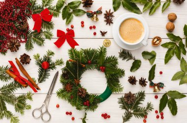 Bir Noel eşyası koleksiyonu ahşap bir masaya yayılır. Yeşillik, fiyonk ve fındık gibi dekorasyonlar var. Bir fincan kahve makasın yanında oturur ve çelenk yapmak için gerekli malzemeleri hazırlar..