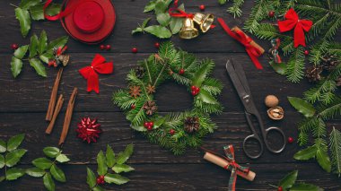 İnsanlar taze yeşillik, kurdele, çan ve süslemeleri kullanarak siyah ahşap bir masa üzerinde tatil çelengi yapıyorlar. Makas ve tarçın çubukları gibi araçlar da mevcuttur..