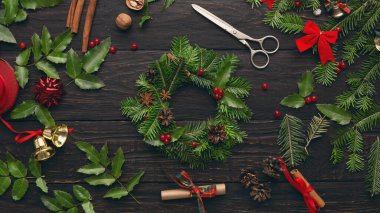 İnsanlar taze yeşiller, kozalaklar ve kırmızı süslemeler kullanarak tatil çelengi yapıyorlar. Makaslar ve kurdeleler yakınlarda ve çeşitli doğal eşyalar koyu ahşap bir masaya yayılmış..