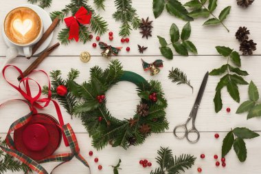 Tatil çelengi yapmak için beyaz ahşap bir masaya çeşitli eşyalar yerleştirilir. Yeşil köknar dalları, kırmızı böğürtlen, kurdele, makas ve bir fincan kahve.