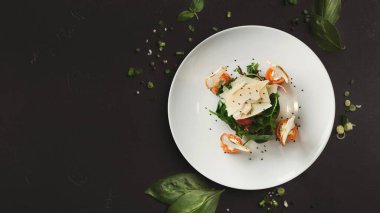 Basit beyaz bir tabakta bir salata seti düzenlenir. Malzemeler arasında sebzeler, dilimlenmiş sebzeler ve süslemeler var. Çanak, etrafa saçılmış otlarla karanlık bir yüzeyde duruyor..