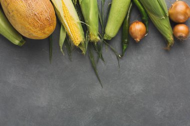 A collection of fresh vegetables is laid out on a dark surface. The set includes corn, beans, onions, and squash. These items can be used for cooking or meal preparation.