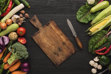 Koyu ahşap bir masadaki ahşap kesme tahtasının etrafında çeşitli taze sebzeler düzenlenir. Tahtaya bir bıçak konur, yemek pişirmeye hazır..