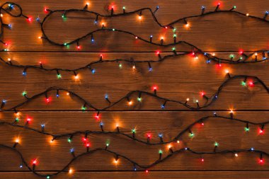 Colorful string lights are spread out in a random pattern on a brown wooden surface. The lights shine in red, blue, green, and yellow. This setup can be used for decoration during winter events.