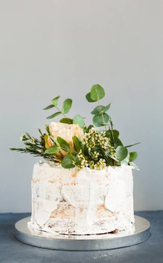 Bir pasta gri arka planda çiçekler ve yeşillikle süslenir. Pasta, beyaz kremalı ve doğal elementli basit bir tasarım içeriyor. Kutlama ya da toplanma için hazır..