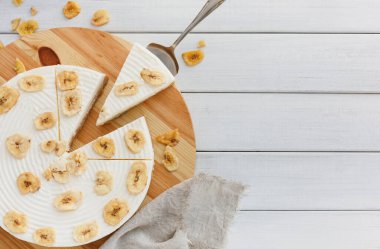 Muzlu bir tatlının birkaç dilimi ahşap bir tahta üzerinde düzenlenir. Tahta beyaz bir masada oturuyor. Kurutulmuş muz parçaları tatlının etrafına detay ekler..