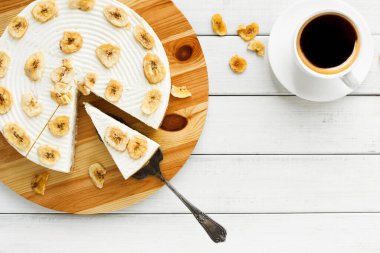 Bir muz kremalı turtası yuvarlak bir tahta üzerine yerleştirilir. Bir dilim kesilir ve yanında servis aracı olur. Beyaz masadaki turtanın yanında bir fincan kahve var..