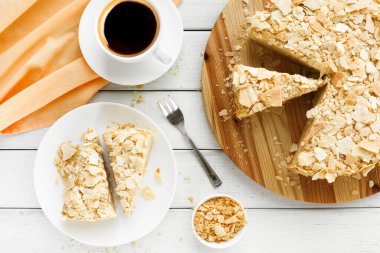 Masada dilimlenmiş yuvarlak bir pasta var. Bir fincan kahvenin yanında bir tabakta iki dilim var. Küçük bir kasede fazladan kek parçaları var. Turuncu kumaş sahneye renk katar.