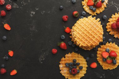 Karanlık bir yüzeyde bir waffle seti düzenlenir. Taze yaban mersini, çilek ve ahududu waffle 'ların etrafına saçılmış renkli bir görüntü oluşturuyor..