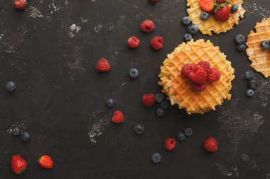 Koyu bir yüzeye yerleştirilmiş waffle 'lar ahududu ve yaban mersinleriyle kaplıdır. Taze çilekler etrafa saçılmış. Bu renkli düzen lezzetli ve davetkar bir sahne yaratıyor..