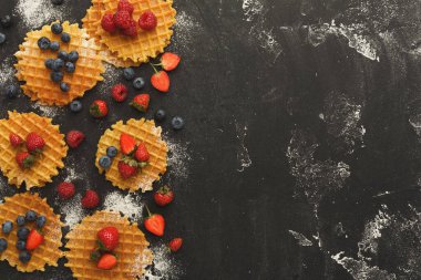 Waffle 'lar siyah bir yüzeye yerleştirilir. Waffle 'ların üzerine farklı meyveler konur. Waffle 'ların etrafına biraz pudra şekeri serpilmiş. Bu kurgu davetkar ve lezzetli görünüyor..