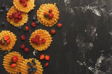 Karanlık bir yüzeyde yuvarlak bir waffle seti düzenlenir. Taze çilek, yaban mersini ve ahududu waffle 'ların üzerine ve etrafına konur. Bu ayar renkli bir gıda görüntüsü gösterir.