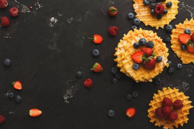 Üzerinde çilek, yaban mersini ve ahududu bulunan dört waffle koyu bir yüzeye yerleştirilir. Bazı böğürtlenler yakınlarda. Bu kurulum kahvaltı sahnesini gösterir.