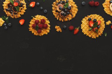 Bir waffle seti mozaik tarzda sergilenir. Her gözlemenin üzerinde farklı meyve ve soslar var. Sahne, koyu bir arkaplan karşısında canlı renkler gösteriyor. Bu kurulum bir yemek sunumu için..