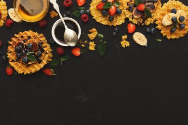 Siyah bir masanın üzerinde bir waffle seti düzenlenir. Her waffle çilek, yabanmersini ve muz gibi meyvelerle kaplıdır. Yakınlarda bir kase şurup ve kaşık da var..