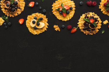 Karanlık bir yüzeyde bir waffle seti bulunur. Her waffle 'da meyve ve çikolata gibi farklı malzemeler var. Sahne çeşitli renk ve dokuları gösteriyor. Waffle 'lar mozaik tarzda düzenlenmiş..