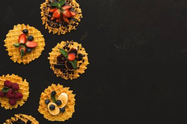 Siyah bir yüzey üzerinde bir waffle seti sergilenir. Her waffle çilek, yabanmersini, muz ve ahududu gibi çeşitli meyvelerle kaplıdır. Düzenleme renkli ve çekici görünüyor..