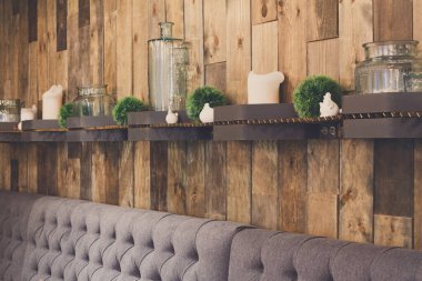 Raflar, kavanozlar, bitkiler ve küçük dekoratif eşyalar ahşap bir duvara dizilmiştir. Aşağıda rahat, gri bir bank var. Burası bir kafede sıcak ve davetkar bir yer..